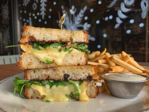 A stacked grilled cheese sandwich with melted cheese and greens, served with fries and a small bowl of dipping sauce.