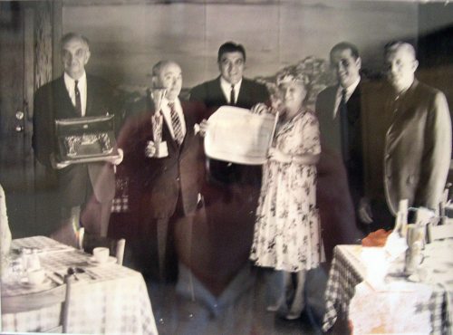 A black-and-white photo of six people, including a woman, posing with awards and trays in a dining setting.