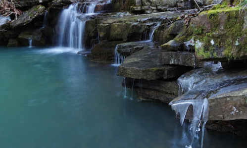 A serene waterfall cascades over rocky ledges, with icicles hanging from the stones above a tranquil blue pool.