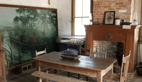 A rustic room with a wooden table, chairs, a brick fireplace, and a large painting of two people in a boat.