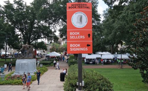 Sign for the Mississippi Book Festival, directing attendees to book sellers and signings, with festival tents in the background.