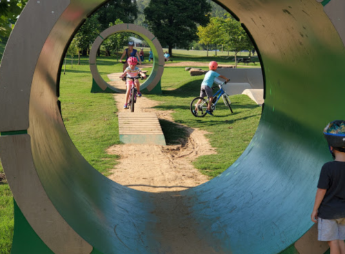 Children riding bikes through circular wooden structures in a park, with green grass and trees in the background.