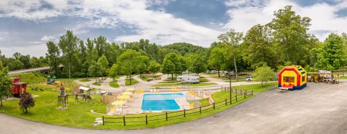 A panoramic view of a campground featuring a pool, playground, and RVs surrounded by lush greenery and trees.