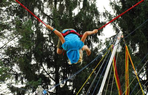 A child in a blue hoodie is upside down on a bungee trampoline, surrounded by tall trees and colorful straps.