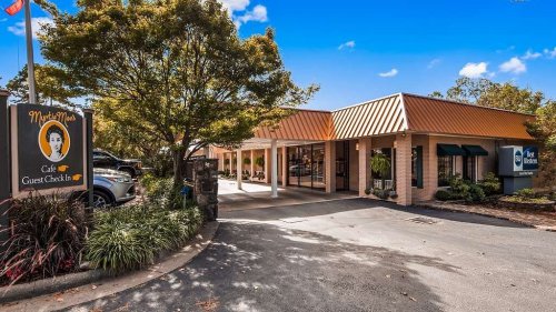 Exterior view of a cafe with a sign, surrounded by trees and parked cars under a clear blue sky.