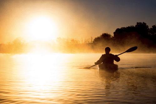 A person kayaking on a misty lake at sunrise, with golden light reflecting on the water.