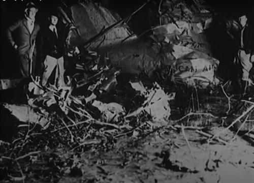 Two men stand near the wreckage of an aircraft, surrounded by debris in a dark, wooded area.