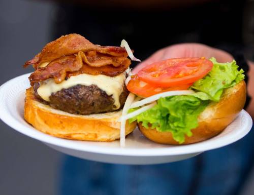 A close-up of a burger with bacon, cheese, lettuce, and tomato on a paper plate, held by a person.