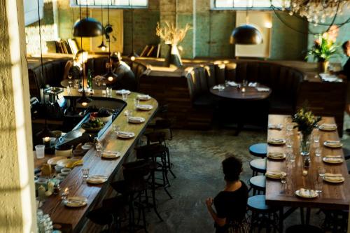 A stylish restaurant interior with a bar, wooden tables, and cozy seating, illuminated by natural light.