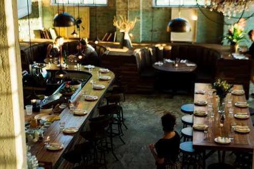 A stylish restaurant interior with a bar, wooden tables, and cozy seating, illuminated by natural light.