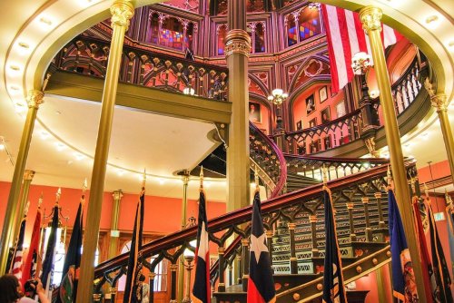 Interior of a grand staircase with ornate railings, surrounded by flags and decorative architecture.