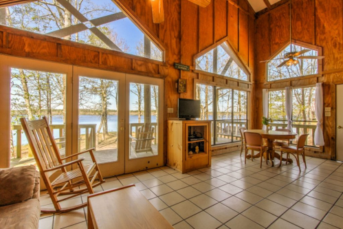 Cozy cabin interior with large windows overlooking a lake, featuring a dining table, rocking chair, and wooden decor.