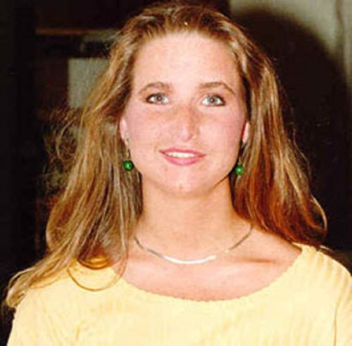 A woman with long hair and green earrings smiles, wearing a yellow top and a silver necklace.