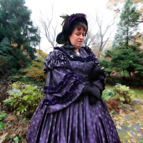 A person in a purple Victorian dress stands outdoors, surrounded by trees and autumn foliage.