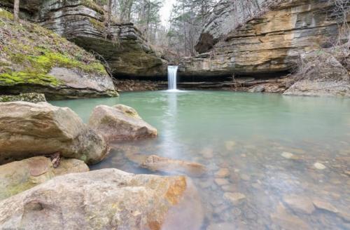 A serene waterfall cascades into a tranquil green pool, surrounded by rocky cliffs and lush vegetation.