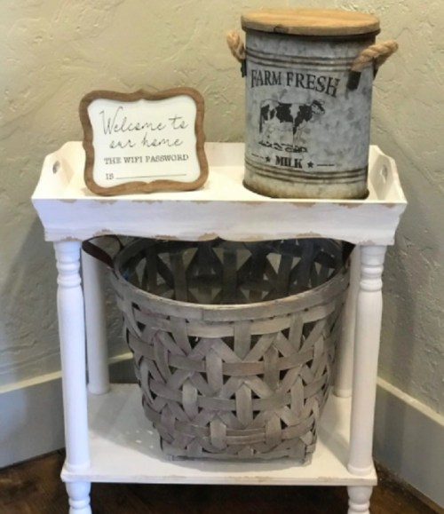 A white table with a sign saying "Welcome to our home," a metal bucket labeled "Farm Fresh Milk," and a woven basket.