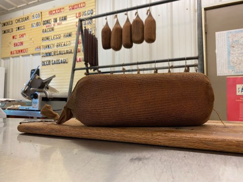 A large, cylindrical sausage wrapped in burlap sits on a wooden cutting board in a butcher shop.