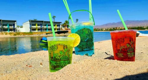 Three colorful drinks with straws sit on a sandy beach, with a calm water backdrop and palm trees in the distance.