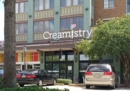 Exterior of Creamistry, a liquid nitrogen ice cream shop, with large windows and a sign, surrounded by trees and parked cars.