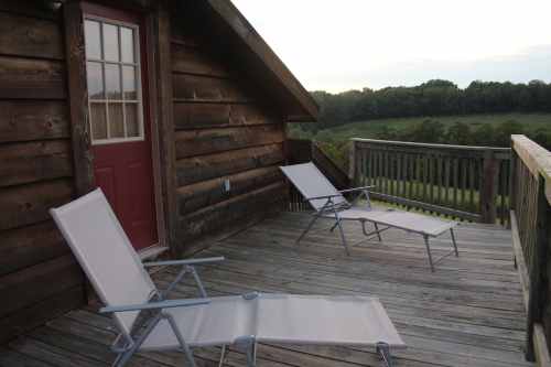 A wooden deck with two lounge chairs and a red door, overlooking a green landscape at sunset.