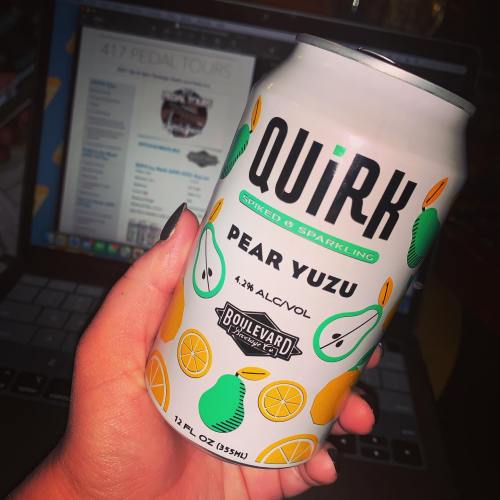 A hand holding a can of Quirk Sparkling Beverage in Pear Yuzu flavor, with a laptop in the background.