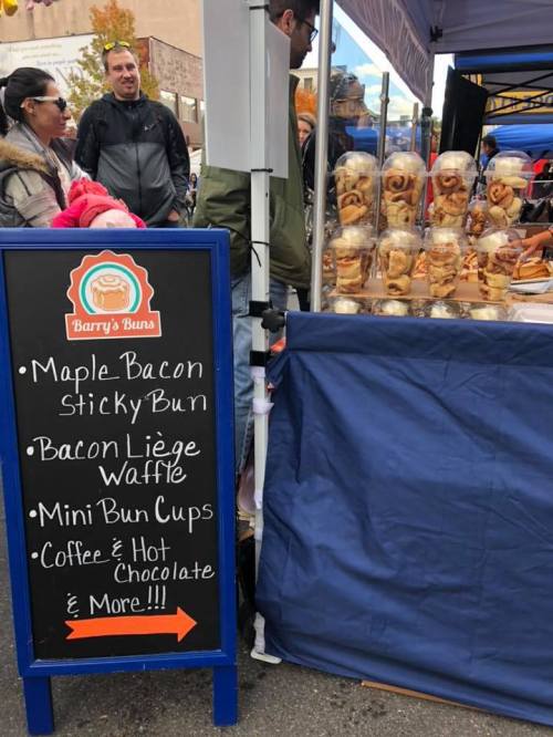 A food stall sign displays menu items like sticky buns, waffles, and drinks, with people browsing nearby.