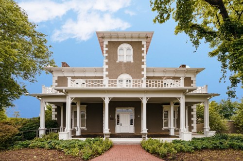 A large, historic house with a porch, white trim, and a distinctive upper window, surrounded by greenery.