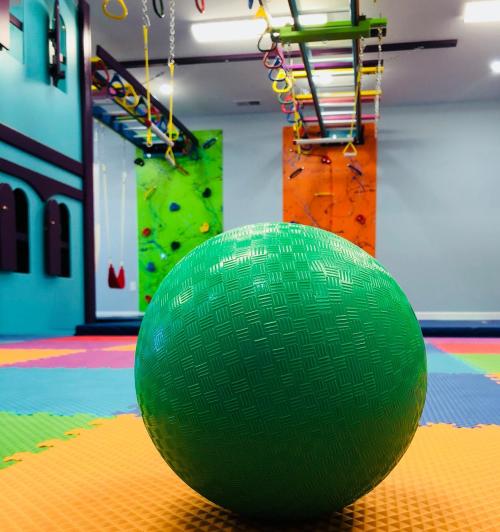 A green ball on a colorful gym floor, with climbing walls and obstacle equipment in the background.