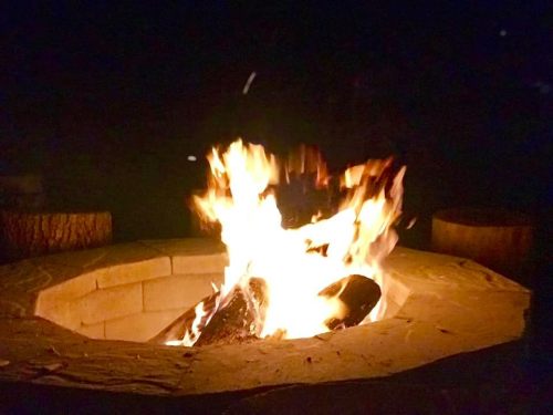 A bright fire burns in a stone fire pit at night, surrounded by wooden seating.