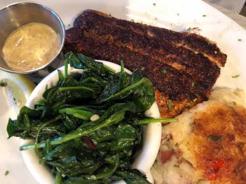 A plate featuring blackened fish, sautéed greens, creamy mashed potatoes, and a small cup of sauce.