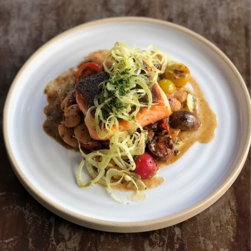A plated dish featuring salmon on a bed of beans and tomatoes, topped with shredded fennel and herbs.