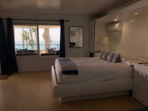 Bright hotel room with a large bed, a window overlooking palm trees, and a balcony with outdoor seating.