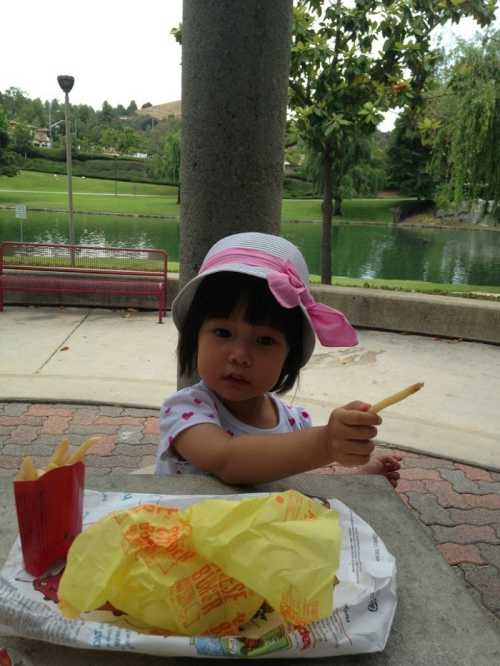 A young child in a hat sits at a table by a lake, holding a French fry with food wrappers nearby.