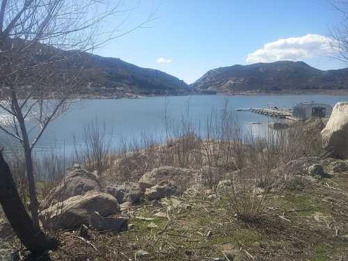 A serene lake surrounded by hills, with a dock and rocky shoreline under a clear blue sky.