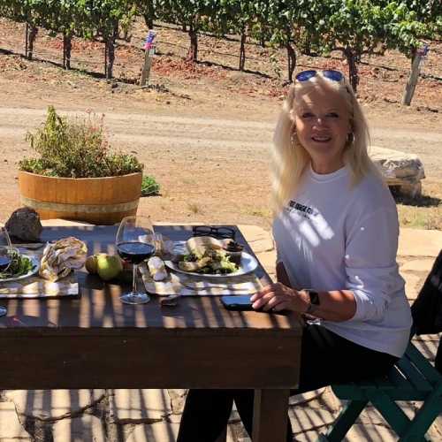 A woman sits at a table in a vineyard, enjoying a meal with wine, surrounded by lush grapevines and a sunny landscape.