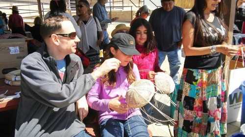 A group of people at a market, with two individuals crafting while others observe and chat in a sunny outdoor setting.