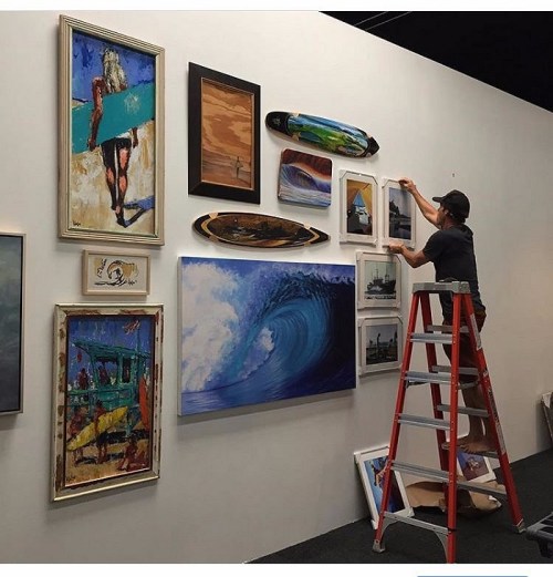 A person on a ladder hangs artwork on a gallery wall featuring various paintings and surfboards.