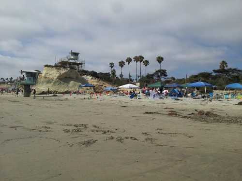 A beach scene with umbrellas, people relaxing, and a lifeguard tower on a rocky shore under a cloudy sky.