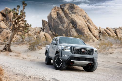 A gray pickup truck parked on a sandy road with large rock formations and a Joshua tree in the background.