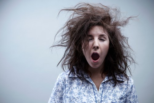 A woman with messy hair yawns, wearing a floral-patterned shirt against a plain background.