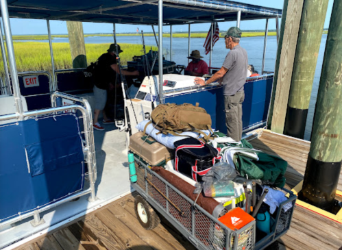 A group of people loading gear onto a boat at a dock, with a scenic marsh view in the background.