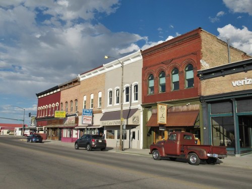 A street view of historic buildings with shops, a vintage red truck parked, and a blue sky with clouds above.