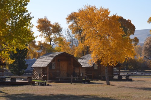 Two rustic log cabins surrounded by vibrant autumn trees with golden leaves under a clear blue sky.