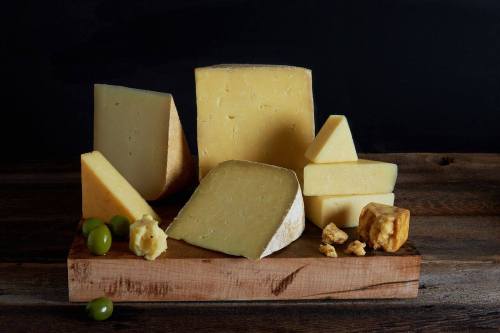 A wooden board displays various types of cheese, including wedges and blocks, alongside green olives and butter.