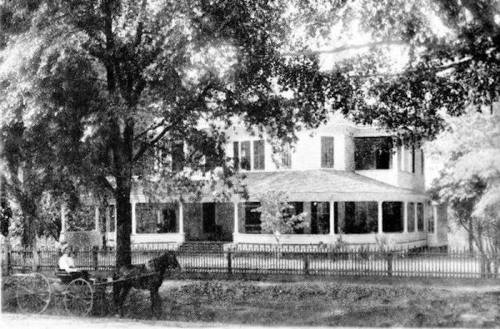 A vintage black-and-white photo of a large house with a horse-drawn carriage in front, surrounded by trees and a fence.