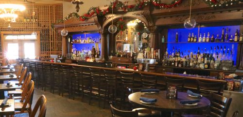A cozy bar with wooden decor, blue-lit shelves of liquor, and festive holiday decorations. Tables are set for dining.