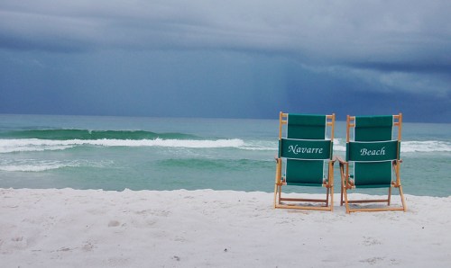 Two green beach chairs labeled "Navarre Beach" sit on a sandy shore, facing a calm ocean under a cloudy sky.