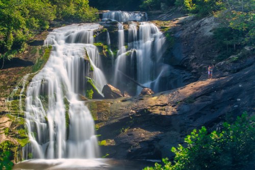 A scenic waterfall cascades over rocky terrain, surrounded by lush greenery, with two people standing nearby.