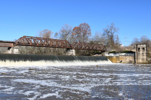 A metal bridge spans a dam with flowing water, surrounded by trees under a clear blue sky.