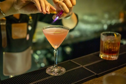 A bartender prepares a pink cocktail with a flame, showcasing a martini glass on a bar counter.